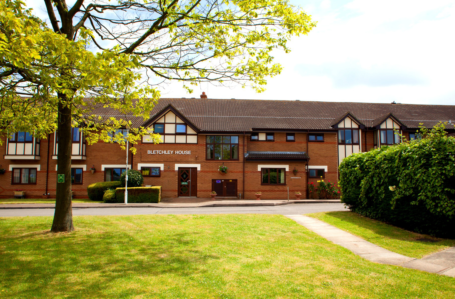 Bletchley House exterior featuring a brick facade, large windows, and a welcoming garden area with a tree and green shrubs.