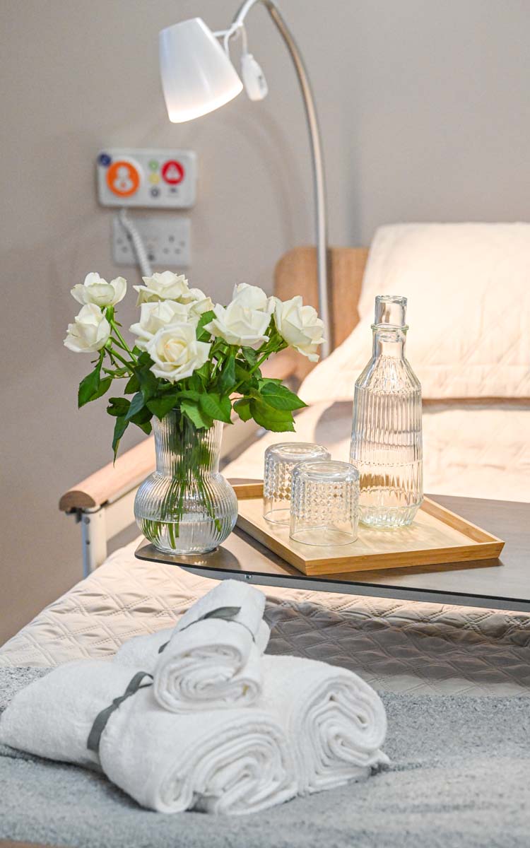 flowers in a vase, a water bottle and two glasses on a table as well as three rolled towels at the end of the bed