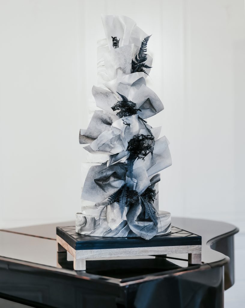 Moderne Hochzeitstorte in Schwarz-Weiß mit dramatischen, abstrakten Zuckerelementen und Farn-Dekor, ein avantgardistisches Kunstwerk von SweetNes Tortendesign, eindrucksvoll inszeniert auf einem schwarzen Flügel in minimalistischem Ambiente.