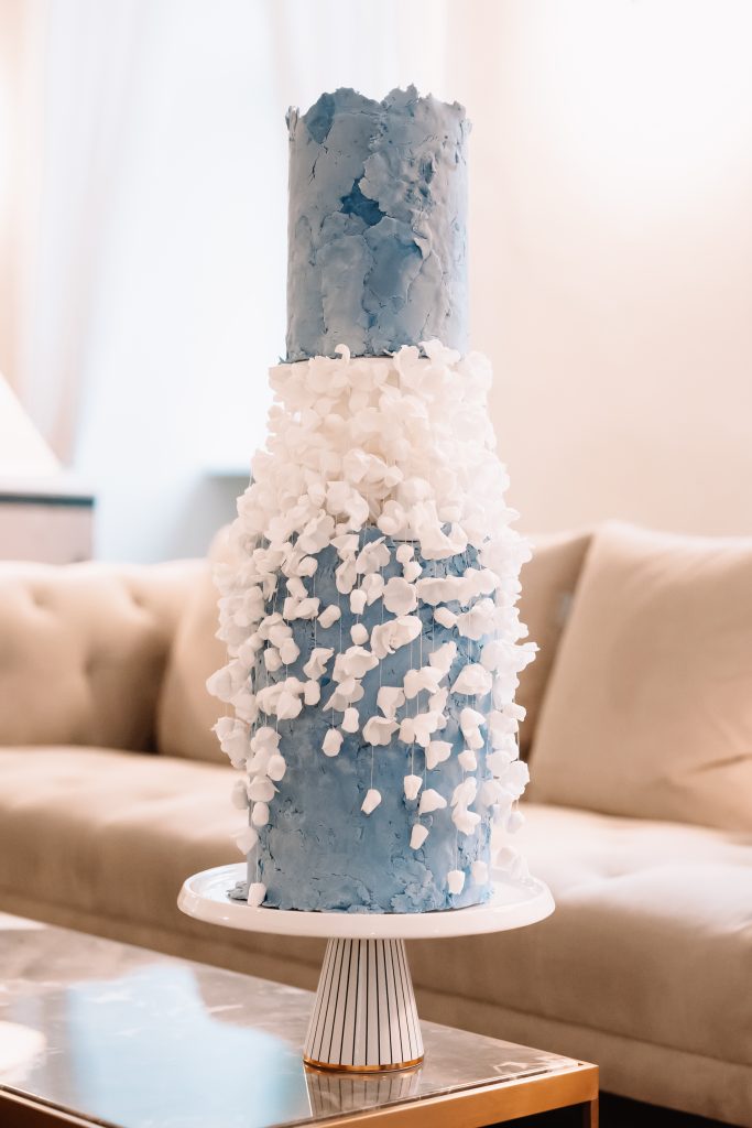 Außergewöhnliche Hochzeitstorte in Blau mit weißen Zuckerblüten in plastischer Struktur – ein kreatives Meisterwerk von SweetNes Tortendesign, präsentiert auf modernem Tortenständer im eleganten Lounge-Ambiente.