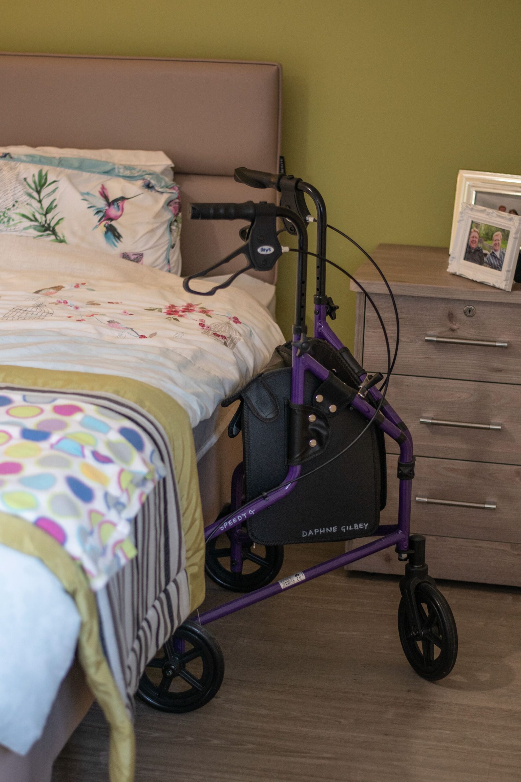 A purple mobility walker beside a bed with floral bedding, next to a chest of drawers and a framed photo on the wall.
