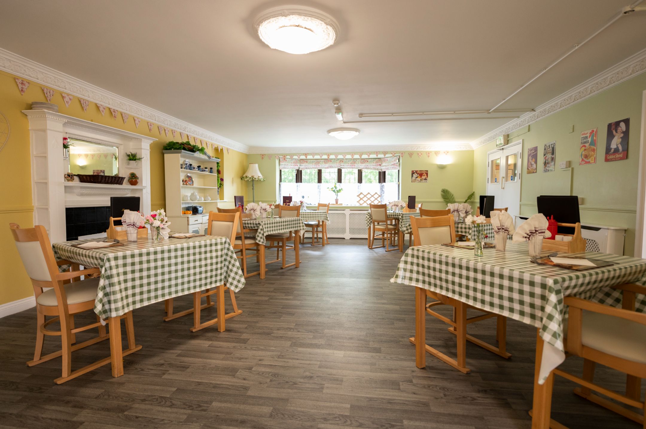 Bright dining area in Tudor Care Home featuring wooden tables with green checkered tablecloths, light-colored walls, and decorative bunting.