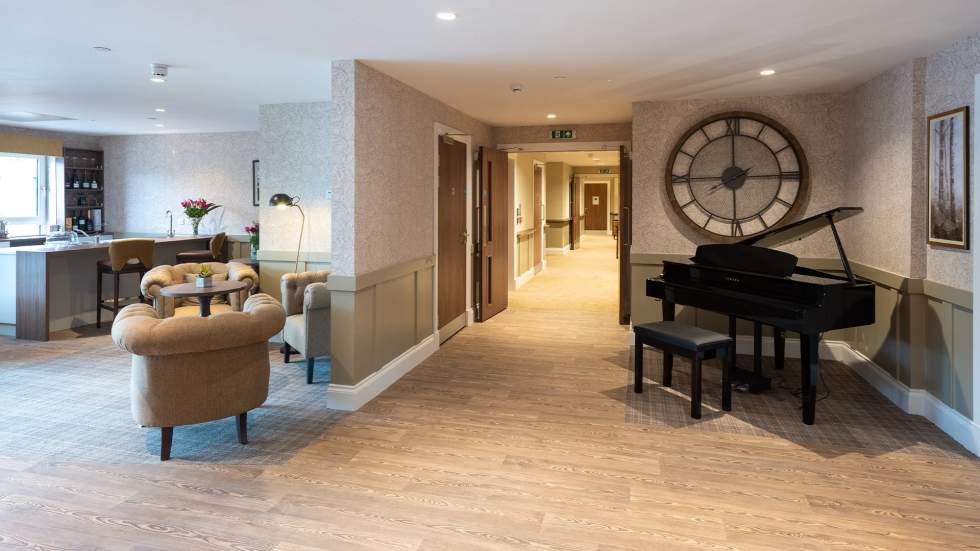 The reception area at the Morar Care Home – Bridge of Weir. There is a small grand piano on the right hand side with a large clock on the wall just behind it. Off to the left is a bar area with a seating area and three armchairs. Behind the bar is a set of shelves with bottles of wine on them. There is a wooden floor and the bar area has a carpet on it.