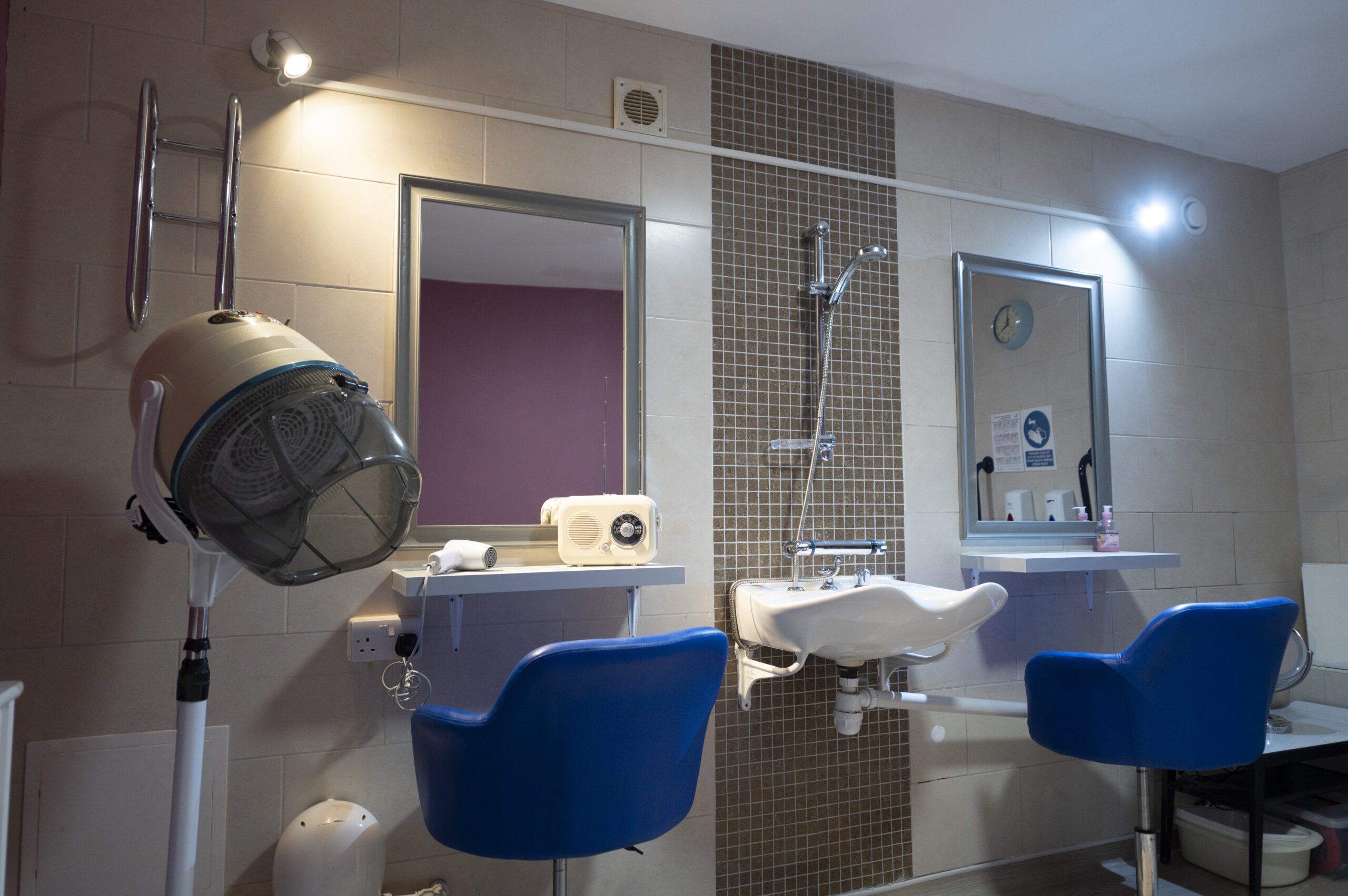 Hairdressing and grooming station at a care home, featuring blue salon chairs, mirrors, a hair dryer, and a sink with modern tiled walls.