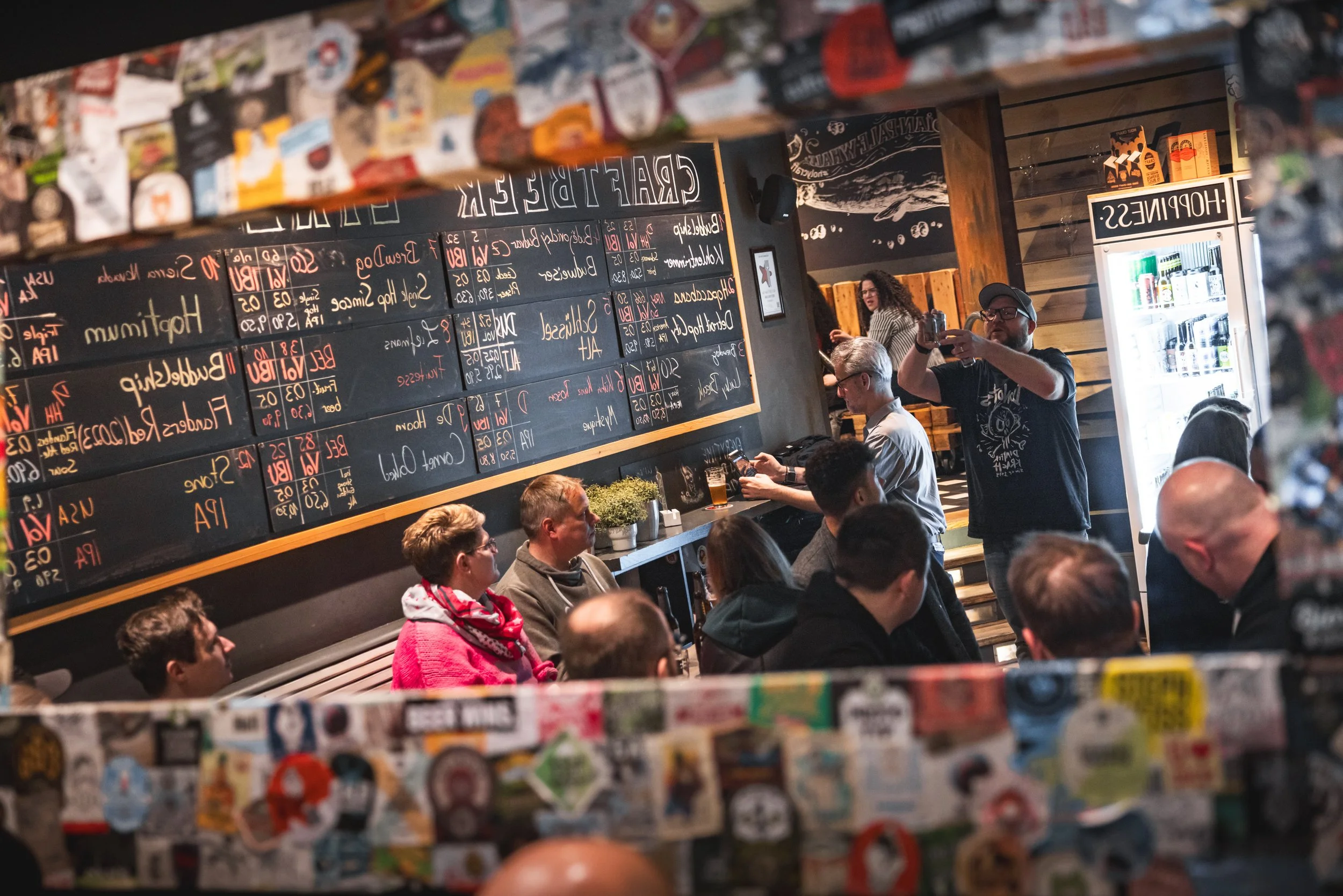 Innenansicht einer Bar mit mehreren Gästen, die an Tischen sitzen und Getränke genießen. Ein Mann macht ein Foto, hinter der Theke ist die Speisekarte auf einer Tafel zu sehen, und im Hintergrund sind eine Zapfanlage und eine Getränkekühlschrank sichtbar.