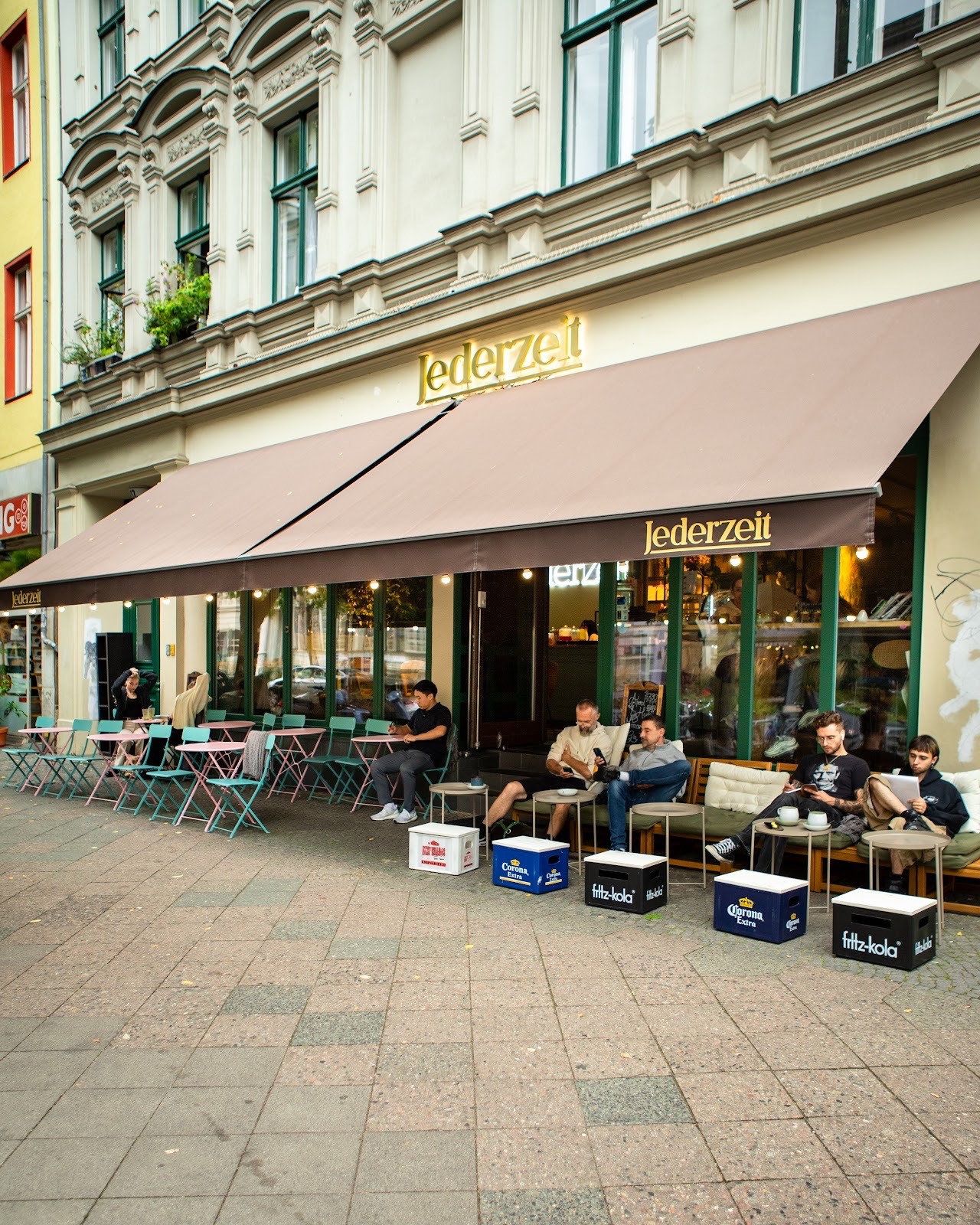 Full exterior view of Jederzeit cafe showing patrons enjoying drinks and food at outdoor tables on the sidewalk.