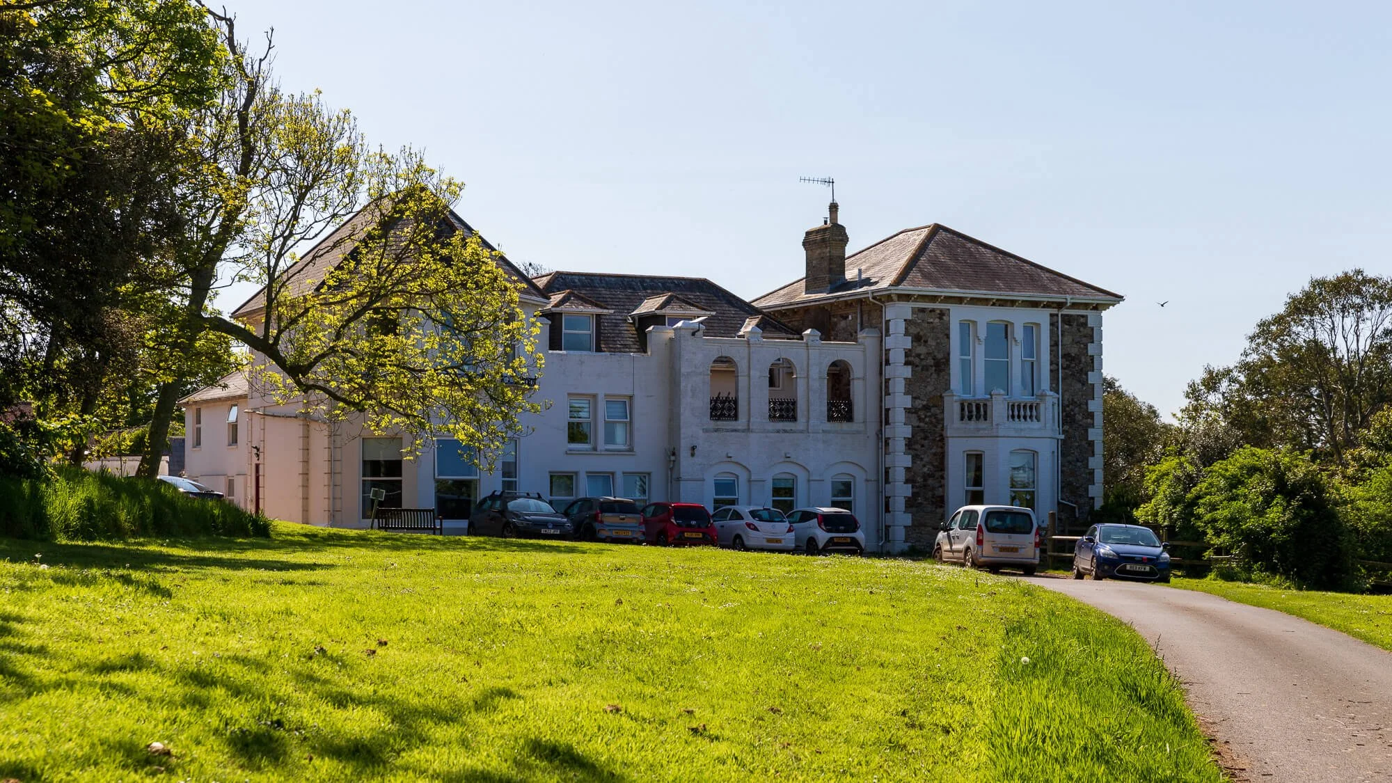 A large, multi-story house with a mix of stone and white exterior walls, surrounded by green trees and a grassy lawn. 