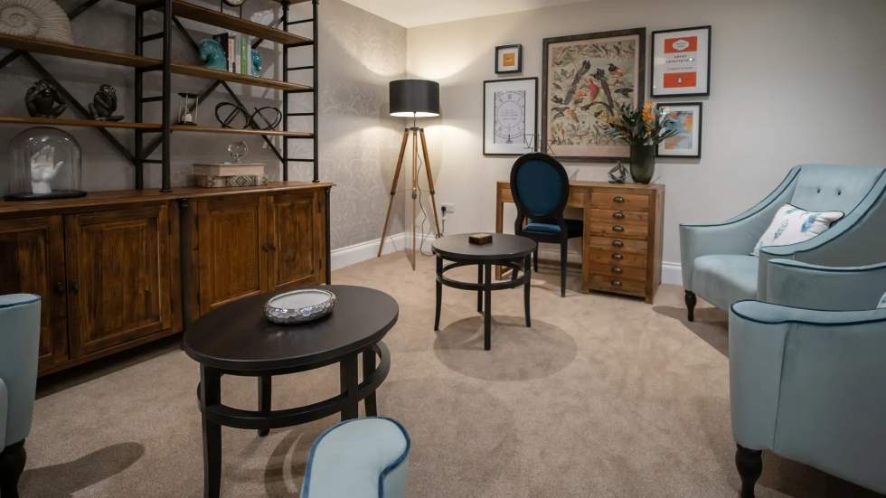 Morar Living Care Homes - Hatfield Manor - a typical quiet room. There is two comfortable armchairs at the edge of the room facing a two circular coffee tables. At one end of the room is a old wooden desk with a chair. In the corner of the room is a tall standing lamp. There is a large sideboard on the wall opposite the chairs with various decorative items on the shelves.