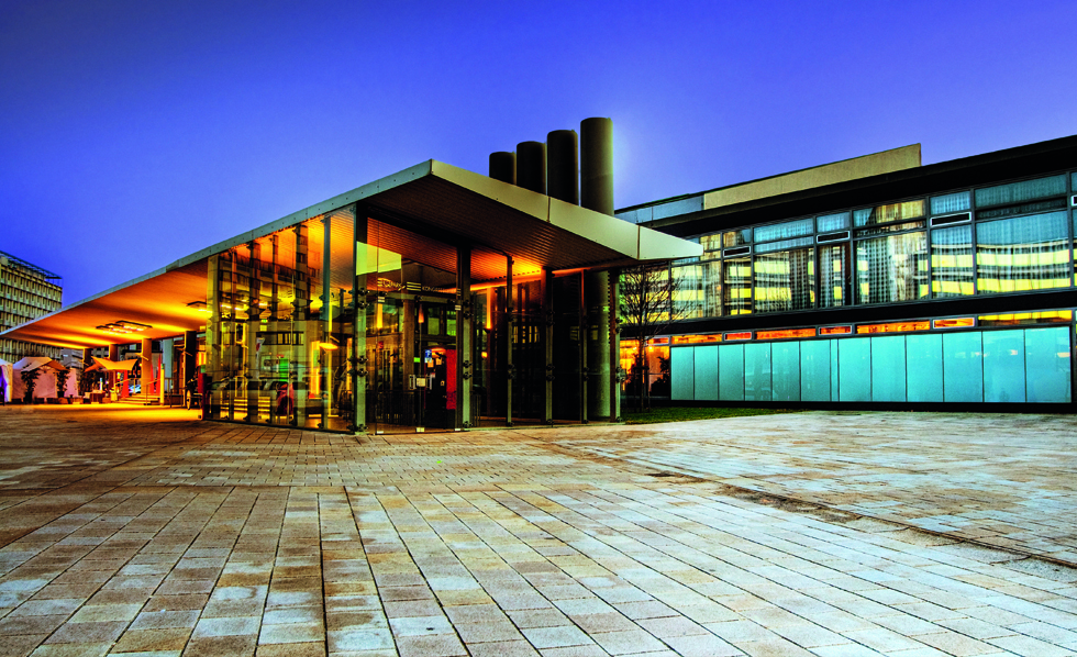 Ein modernes Gebäude mit großen Glaswänden und einem flachen, überhängenden Dach, das in der Abenddämmerung von warmen Lichtern beleuchtet wird. Der gepflasterte Hof davor ist leer, und der Himmel ist tiefblau.